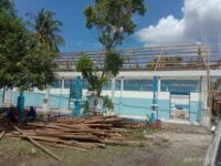Gambar menunjukkan proyek konstruksi di mana konsultan menggunakan kayu lokal kelas 3 untuk memasang seng, menimbulkan perdebatan tentang kualitas dan keamanan material yang digunakan